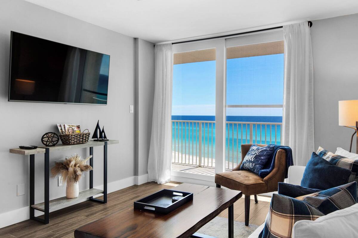 A modern living room with a large TV, a coffee table, a sofa with patterned cushions, and a view of the ocean through large windows.