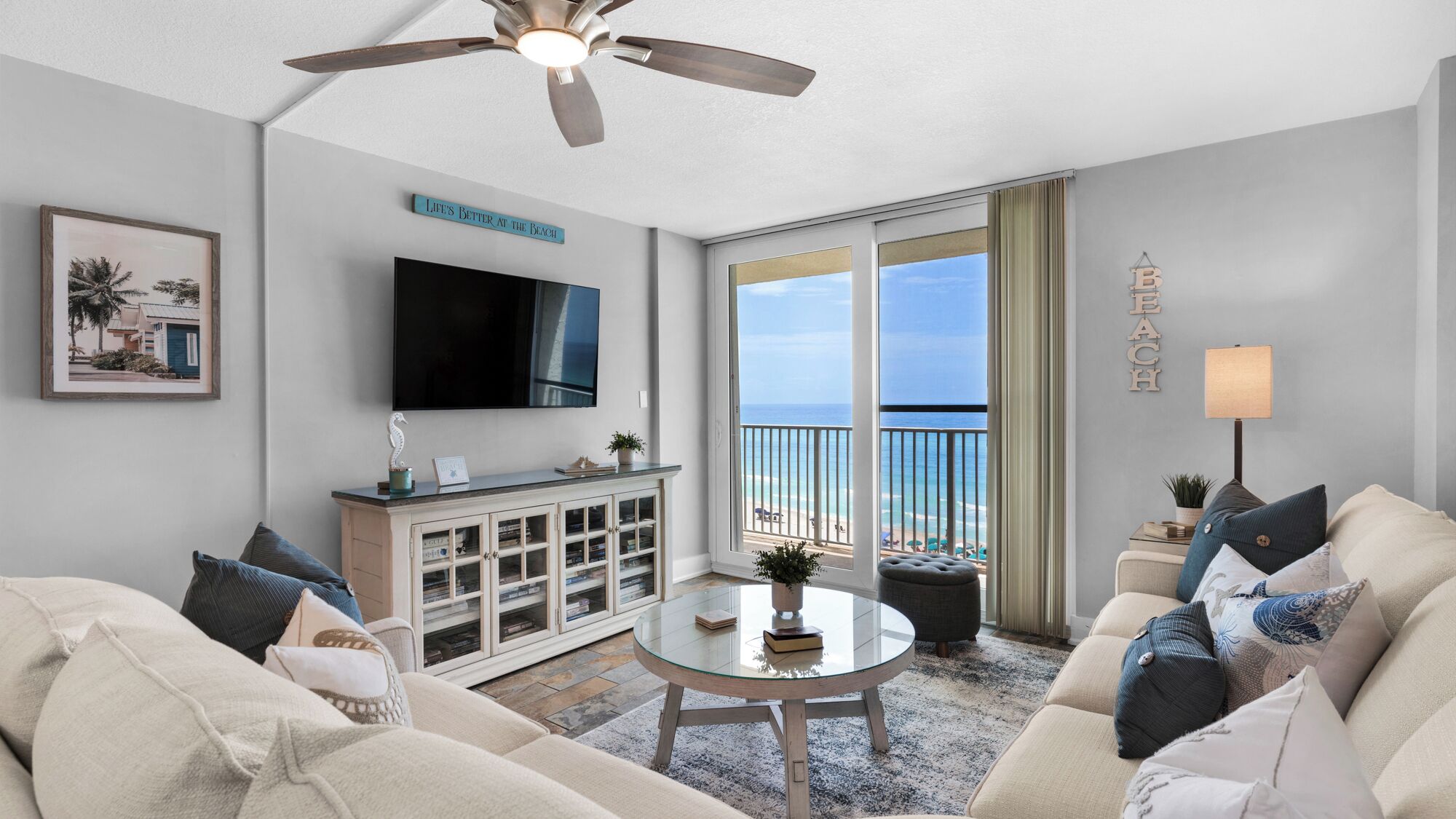 A living room with a TV, a glass coffee table, a sofa, and a view of the beach.