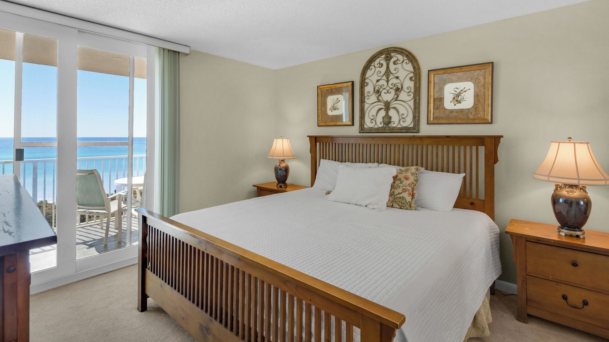 A hotel room with a large bed, two bedside tables, a wooden headboard, and a balcony with a view of the ocean.