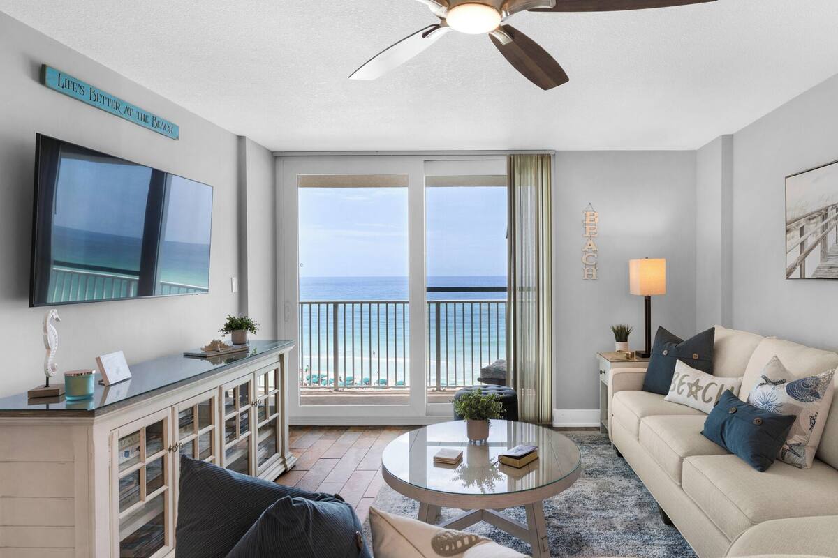 Una sala de estar con un televisor grande, un ventilador de techo, un sofá, una mesa de centro y vista a la playa a través de puertas corredizas de vidrio.