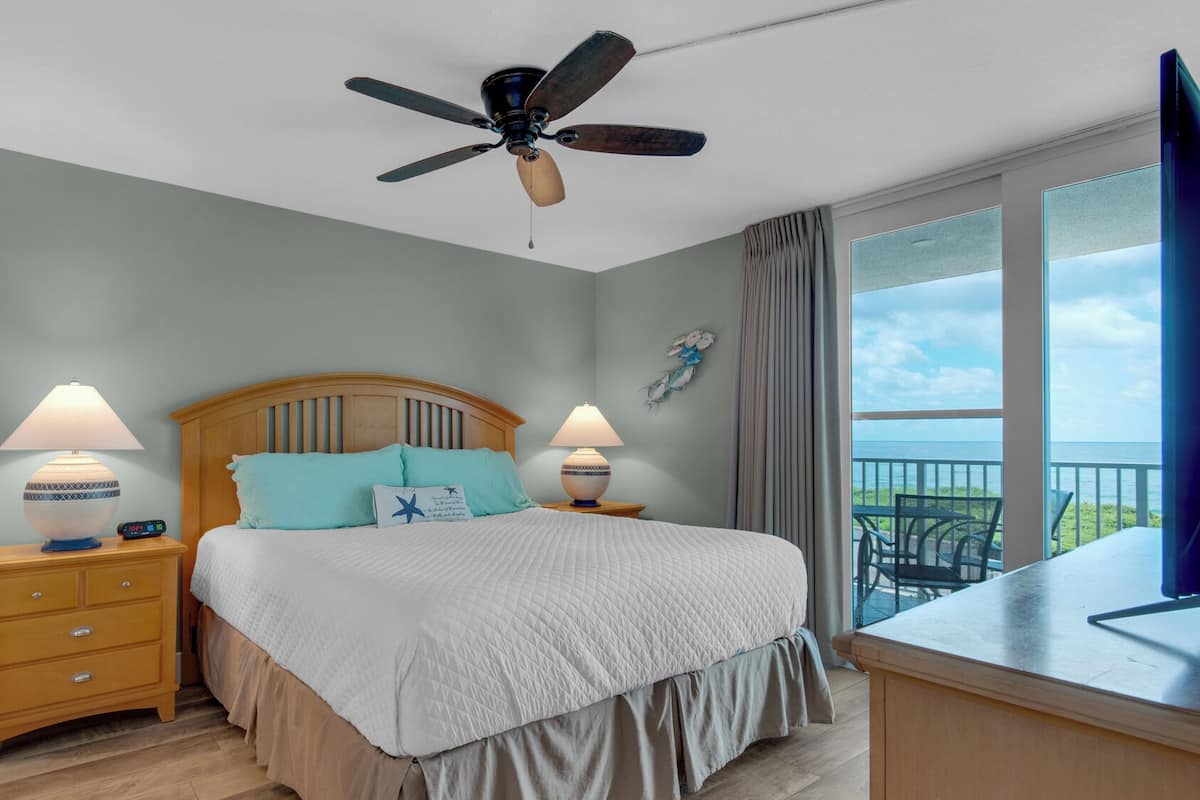 A bedroom with a bed, two bedside tables with lamps, a ceiling fan, a view of the ocean through a balcony, and a small desk with a chair.
