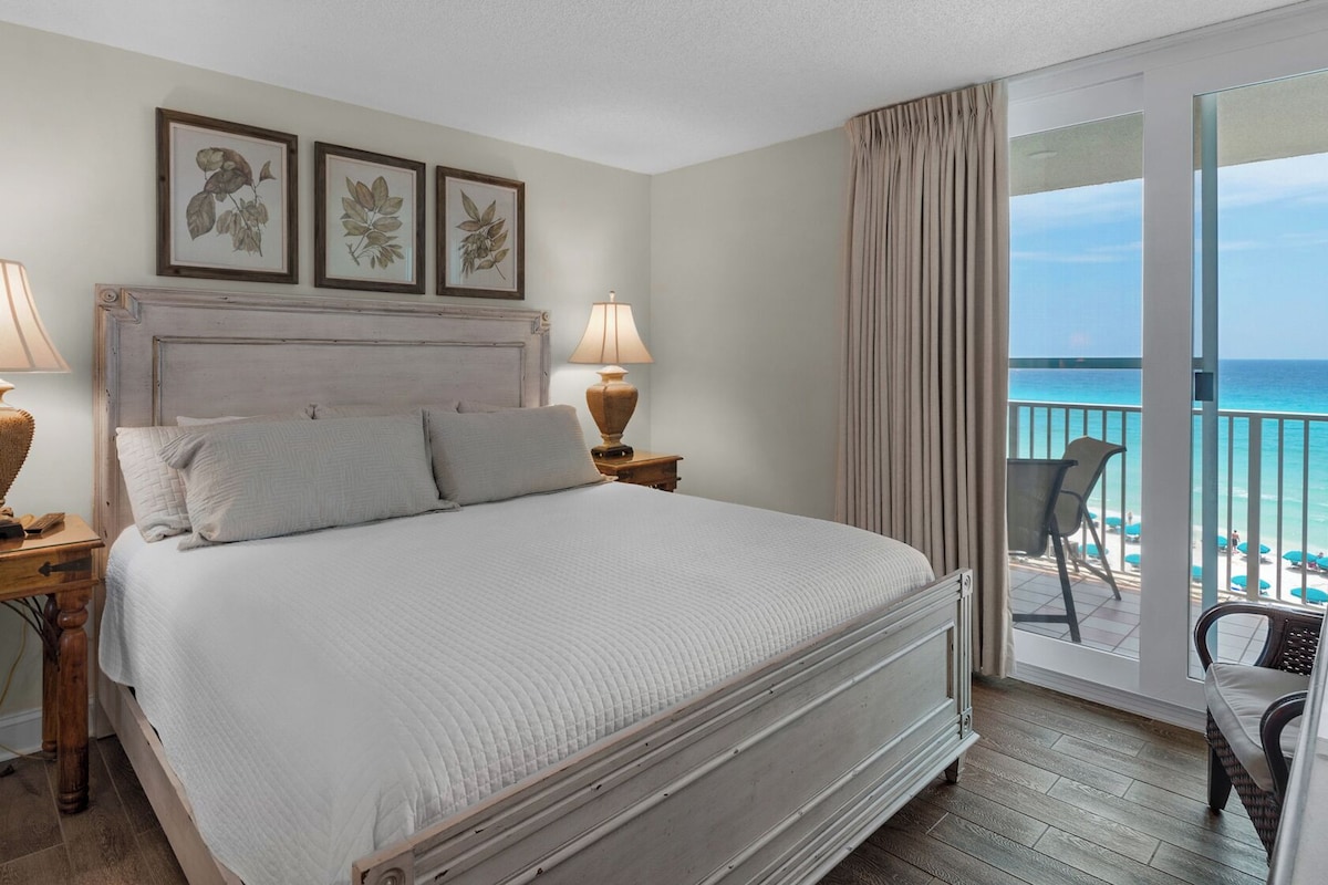 Habitación de hotel con una cama grande, dos lámparas de noche, vista a la playa y un balcón con sillas.