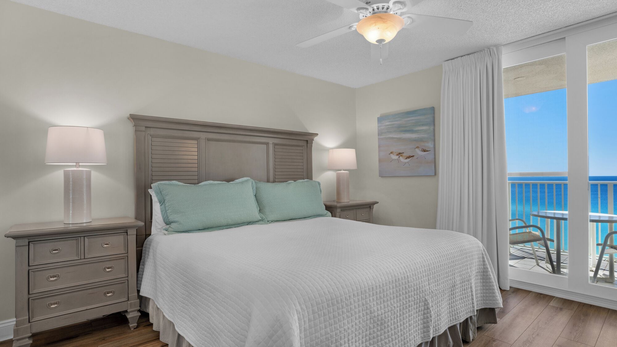 A bedroom with a bed, two bedside lamps, a ceiling fan, and a view of the ocean through a balcony.