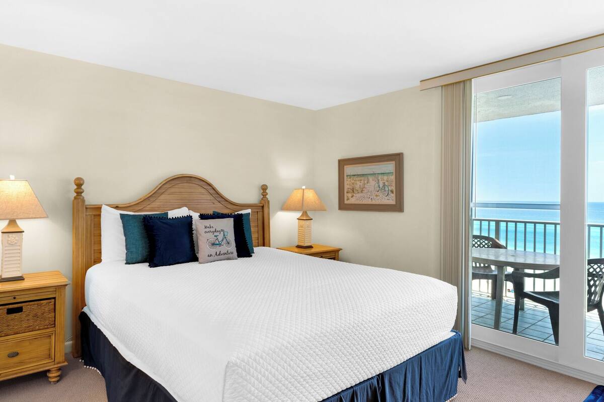 A hotel room with a large bed, two bedside tables with lamps, a view of the ocean through a sliding glass door, and a painting on the wall.