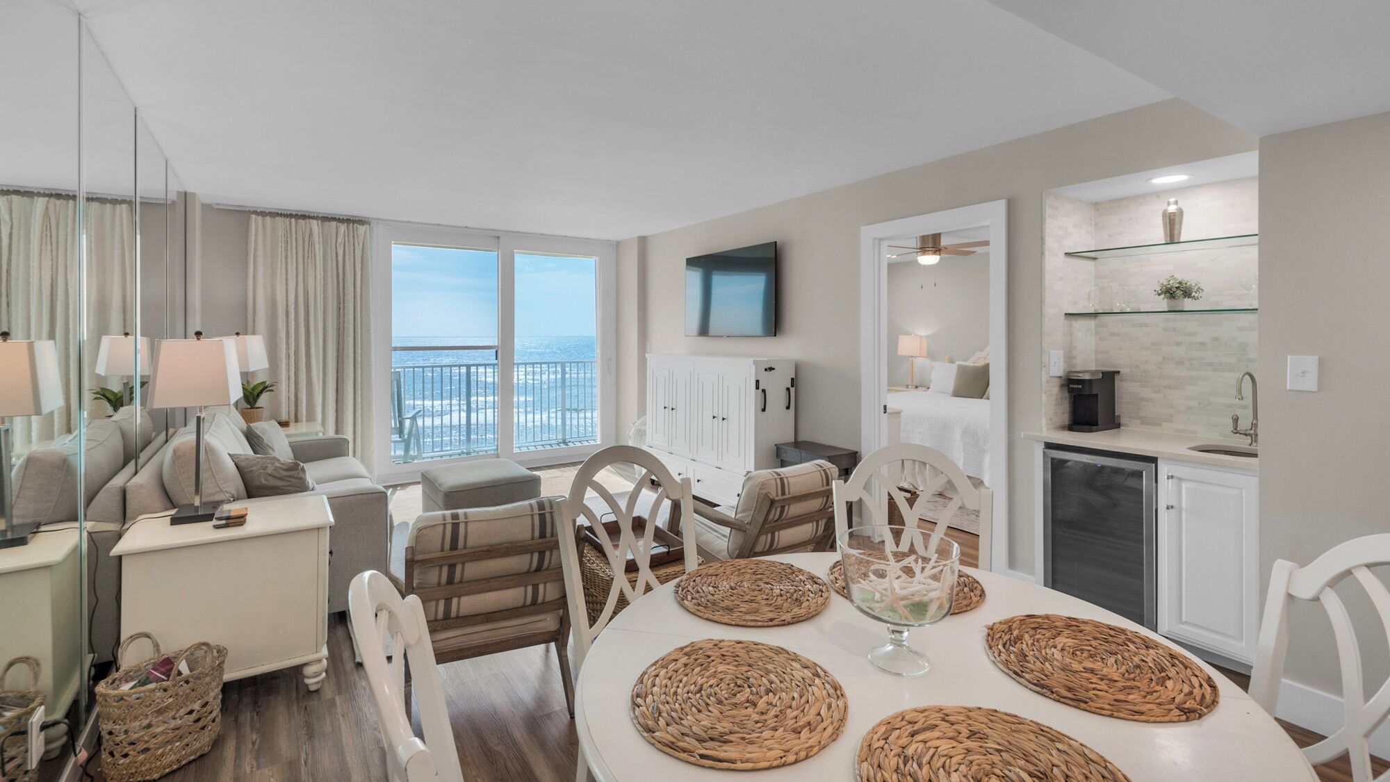 A modern living room with a large window, a dining area with woven placemats, and a kitchen with a glass shelf.