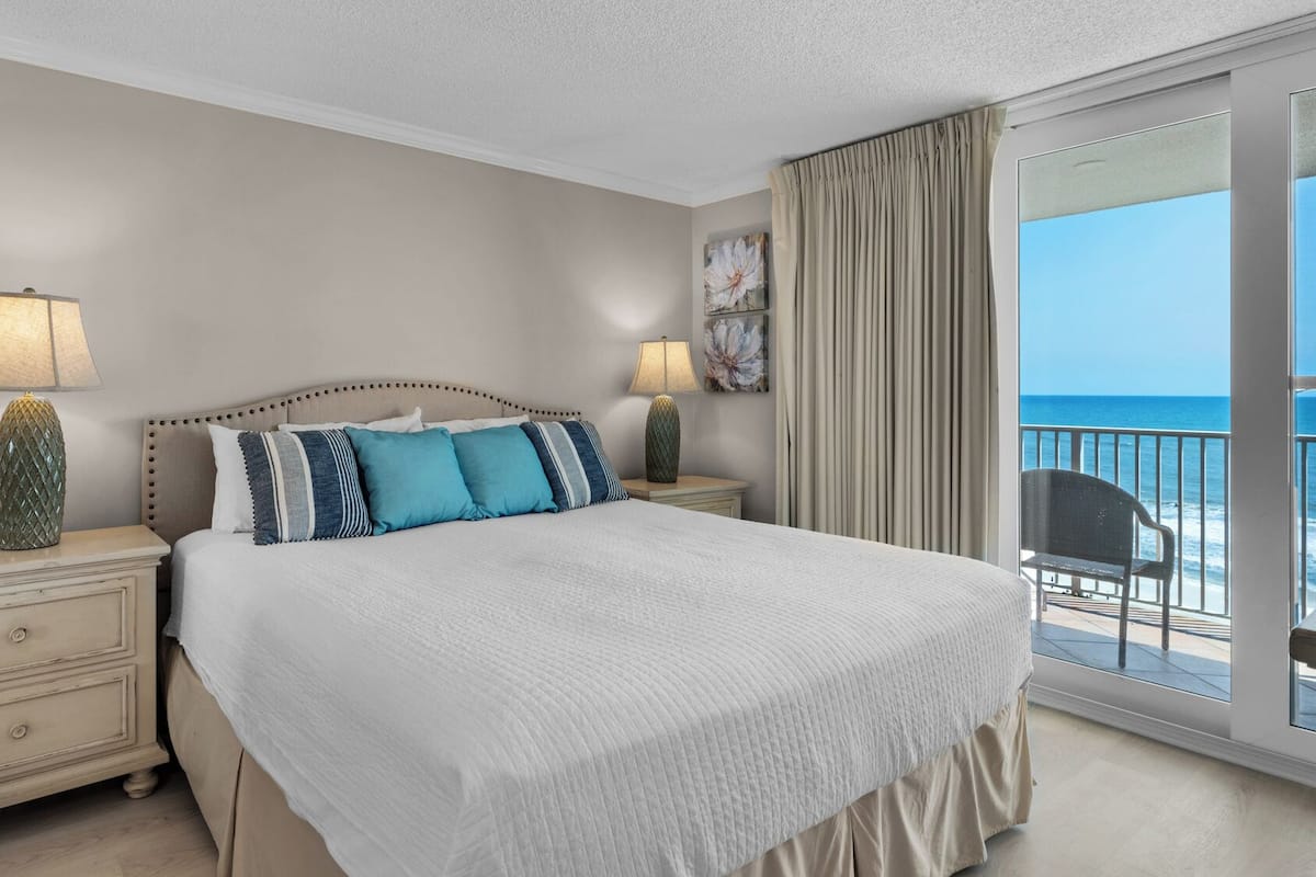 A bedroom with a large bed, two bedside tables with lamps, a nightstand, a view of the ocean through a balcony.
