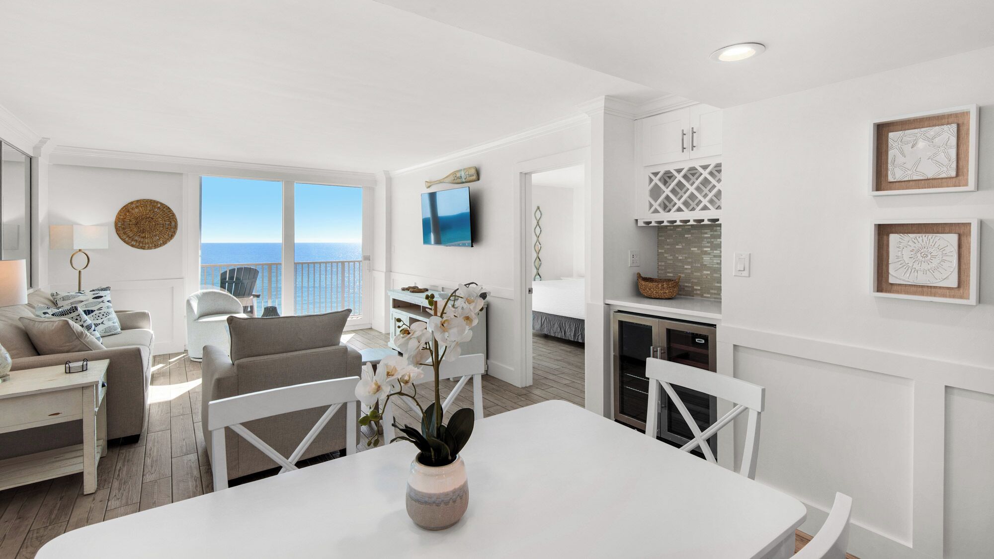 A modern living room with a white sofa, a dining table with chairs, a TV mounted on the wall, and a view of the ocean through large windows.