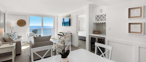 A modern living room with a white sofa, a dining table with chairs, a TV mounted on the wall, and a view of the ocean through large windows.