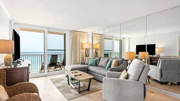 Een moderne woonkamer met een grote bank, fauteuils, een salontafel en een televisie. Er is een balkon met uitzicht op de oceaan.