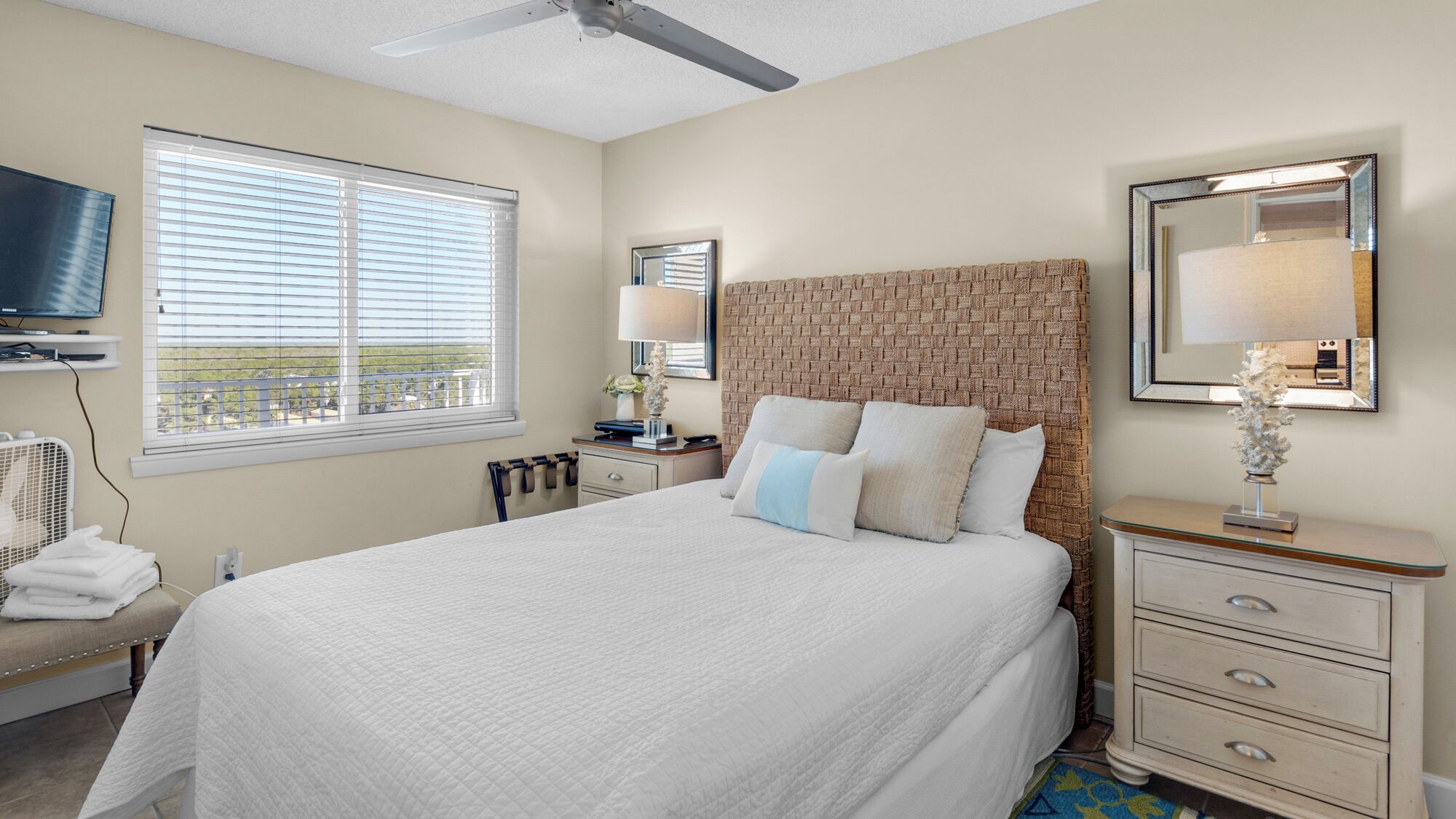 A hotel room with a large bed, a flat-screen TV mounted on the wall, a window with blinds, a ceiling fan, and a nightstand with a lamp.