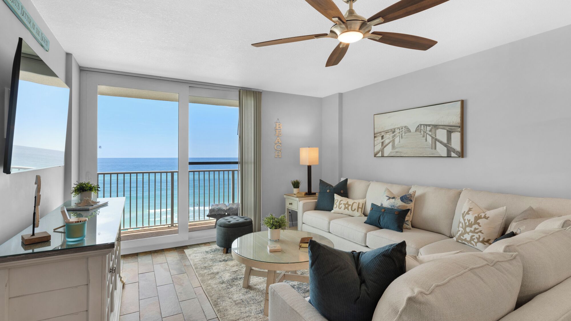 Ein Wohnzimmer mit Blick auf den Strand, einem Deckenventilator, einem Glastisch und einer Couch mit dekorativen Kissen.