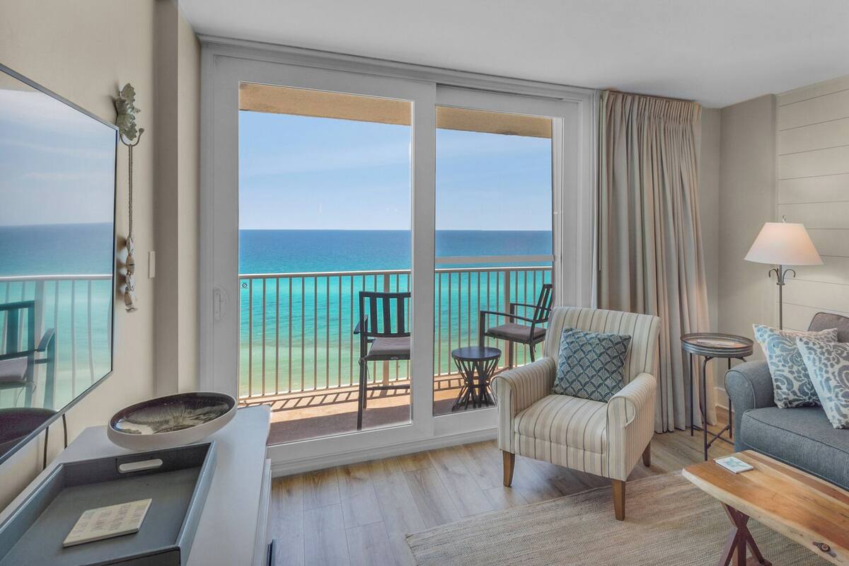A room with a balcony overlooking the ocean, a sofa, a chair, a small table, and a lamp.