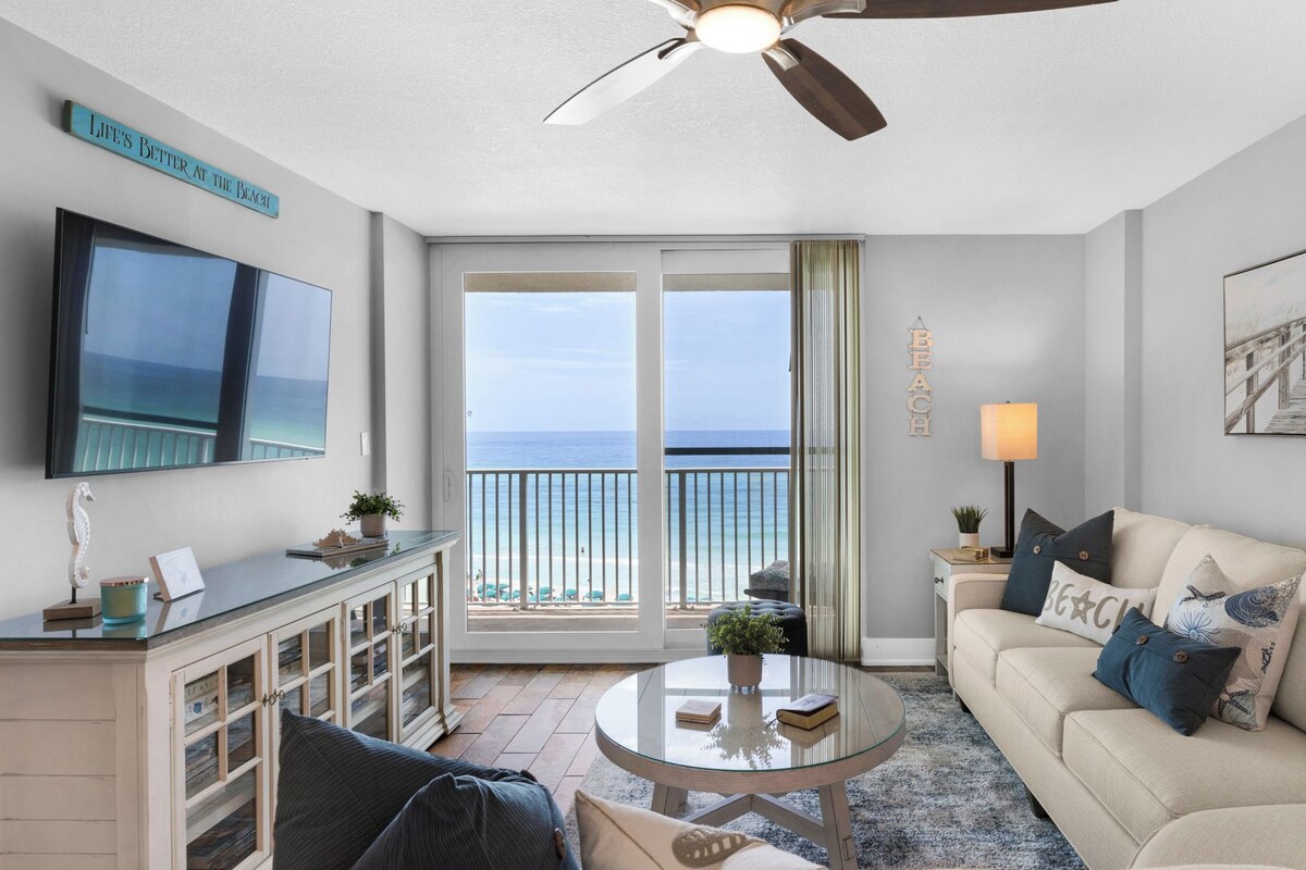 Una sala de estar con un televisor grande, un ventilador de techo, un sofá, una mesa de centro y vista a la playa a través de puertas corredizas de vidrio.