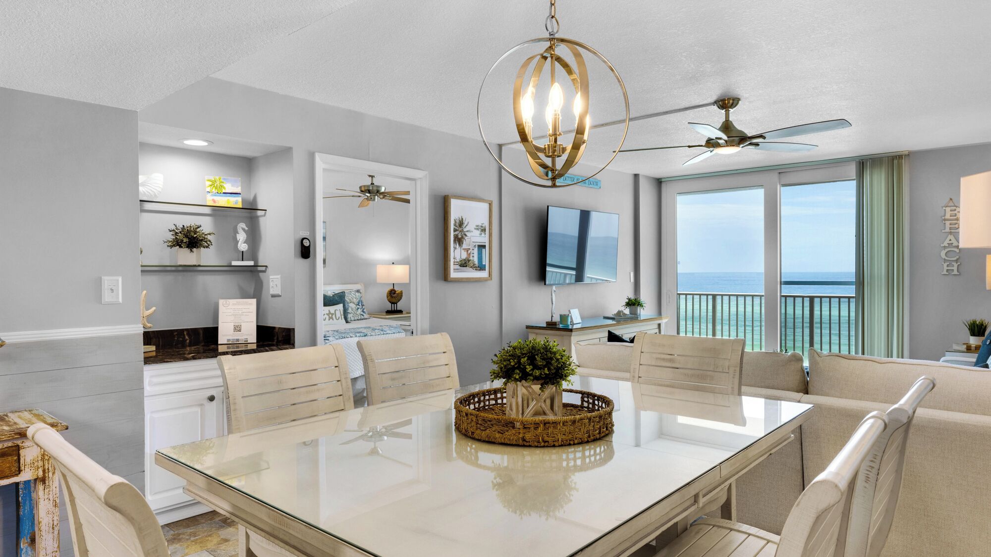 A modern dining area with a glass table, white chairs, a chandelier, and a ceiling fan.