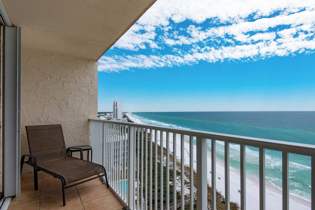 A balcony with a view of the ocean and beach, a chair, and a railing.