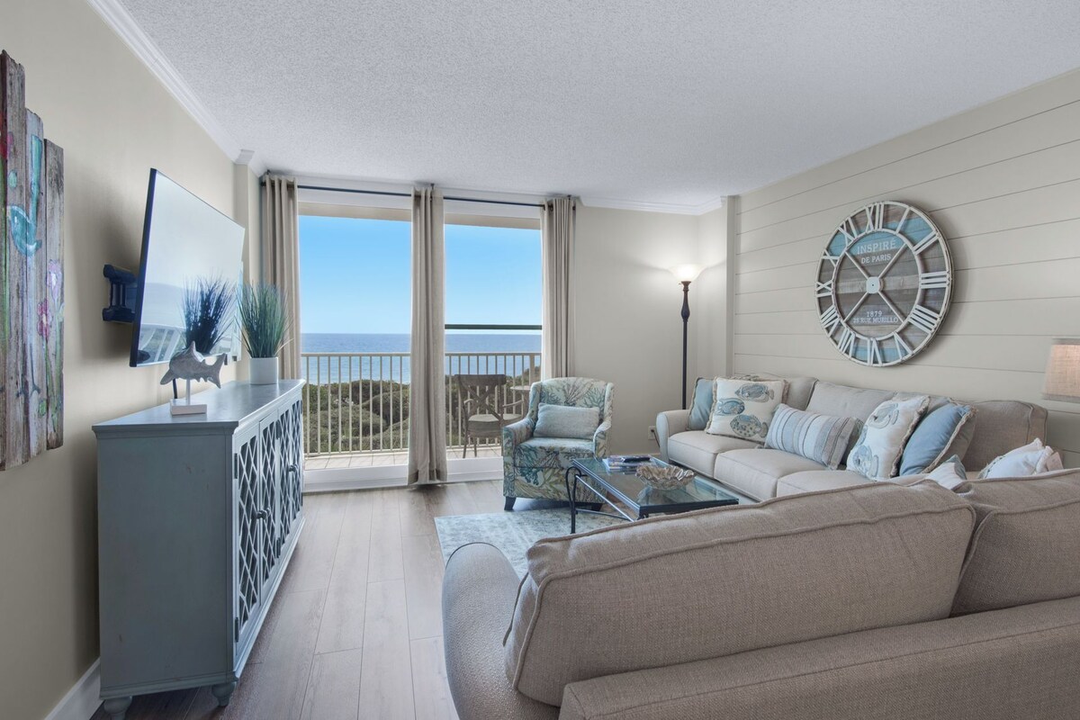 A spacious living room with a balcony, a large wall clock, and a view of the sea.