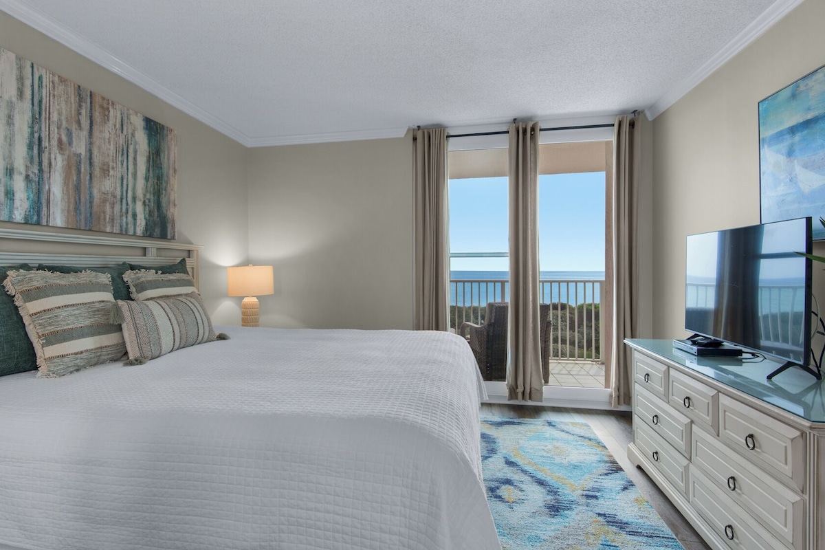 Una habitación de hotel con una cama grande, televisor, escritorio con planta y balcón con vistas.