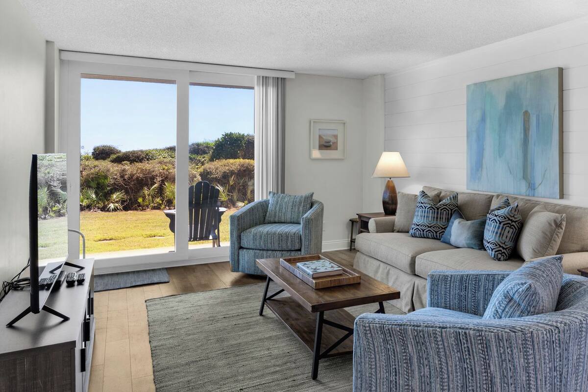 A living room with a sofa, two armchairs, a coffee table, and a TV. There is a sliding glass door leading to a garden.