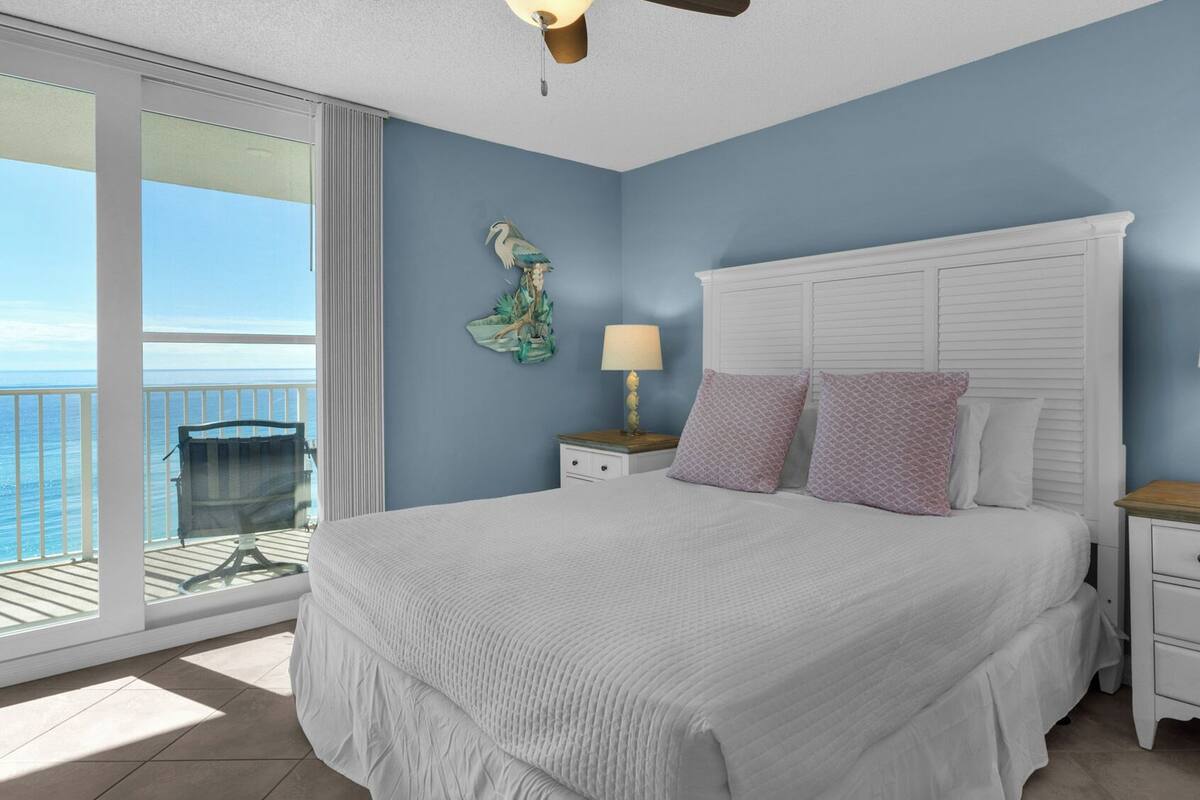 A bedroom with a large bed, two bedside tables with lamps, a nightstand, a decorative wall piece, and a view of the ocean through a sliding glass door.