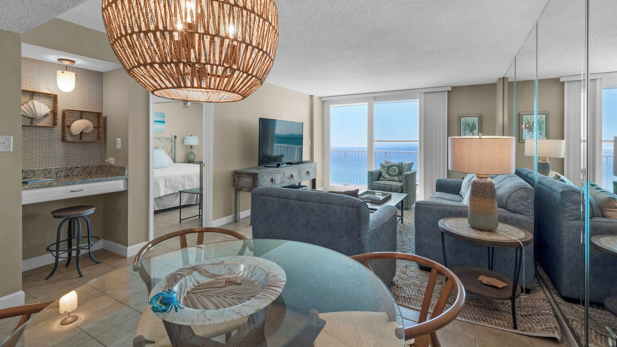 A modern living room with a glass dining table, a sofa, a TV, and a large window with a view of the ocean.
