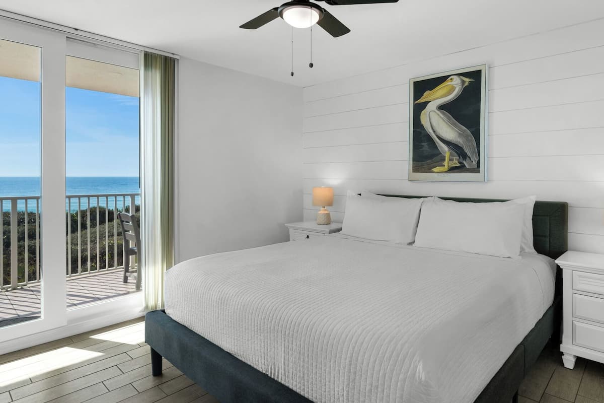 A bedroom with a large bed, two bedside tables with lamps, a ceiling fan, and a balcony with a view of the ocean.