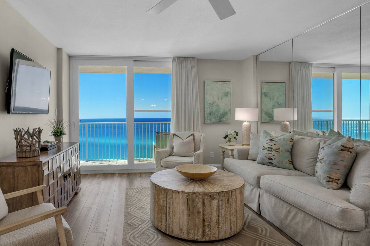 A spacious living room with a large window offering a view of the ocean, a round wooden coffee table, a beige sofa, and a wooden chair.