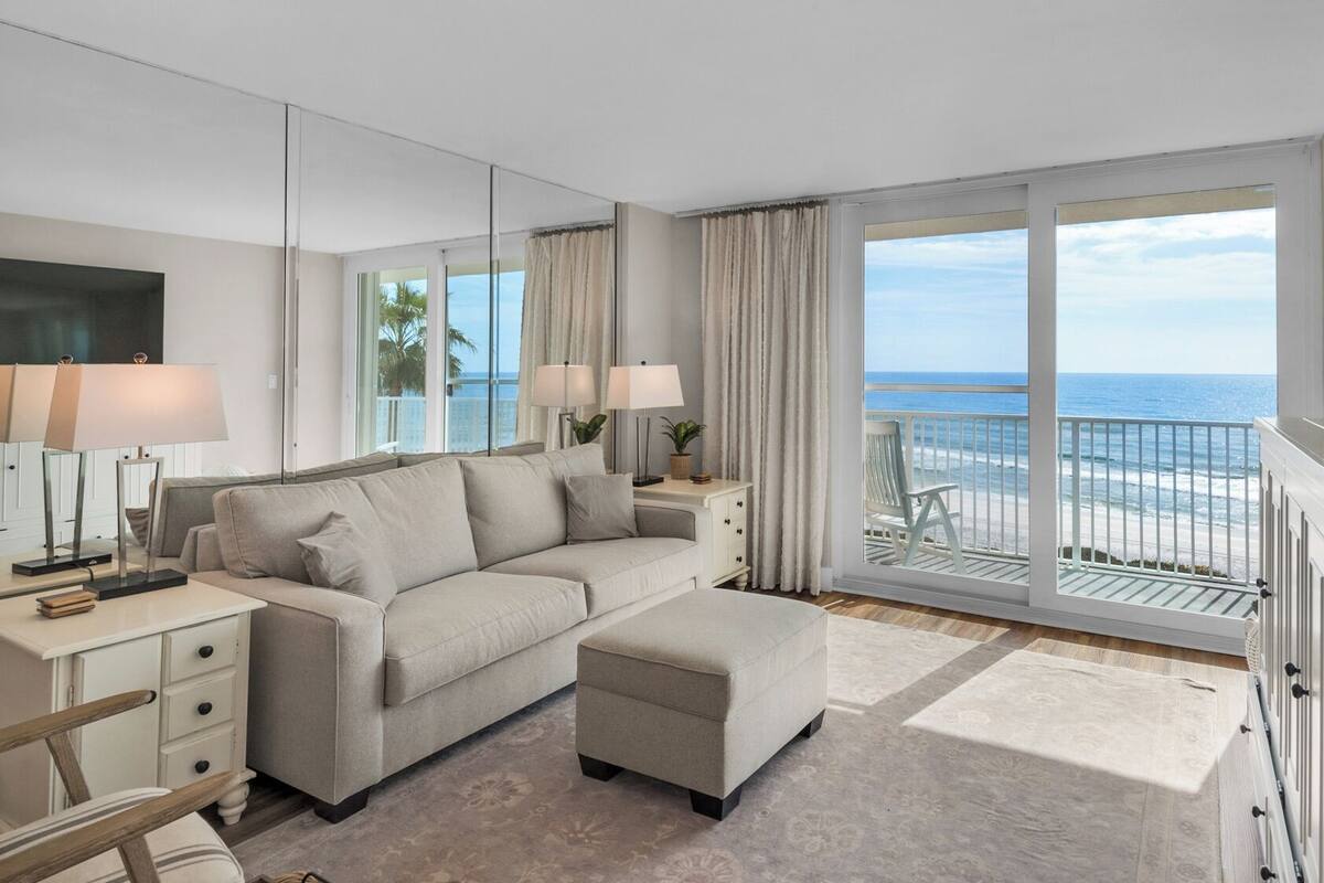 A modern hotel room with a large sofa, a small ottoman, a side table with lamps, a flat-screen TV, and a balcony with a view of the ocean.