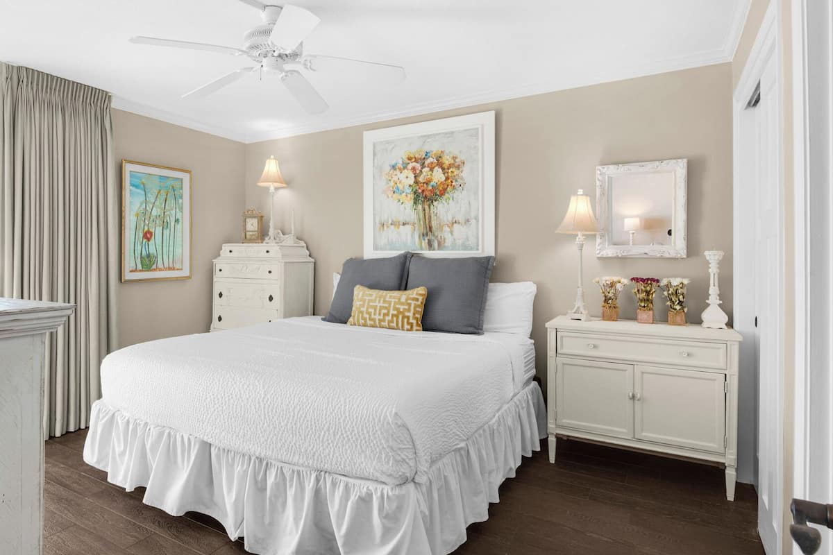 A bedroom with a large bed, a dresser, a nightstand, a ceiling fan, and a window with curtains.