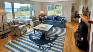 A spacious living room with a view of the outdoors, a fireplace, and a striped rug. - Hard Aground - Ocean View Cottage in Southwest Harbor! Pet Friendly!!!  (Southwest Harbor)