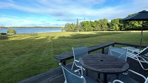 A deck with a round table and chairs, overlooking a grassy area and a body of water. - Hard Aground - Ocean View Cottage in Southwest Harbor! Pet Friendly!!!  (Southwest Harbor)