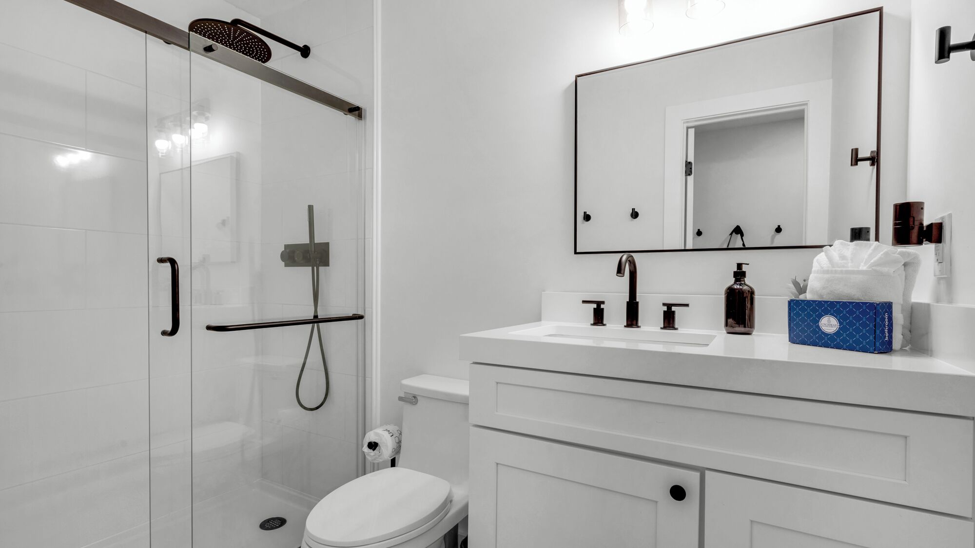 Une salle de bain moderne avec une douche à parois de verre, des toilettes blanches, un meuble-lavabo avec un miroir et un distributeur de serviettes.