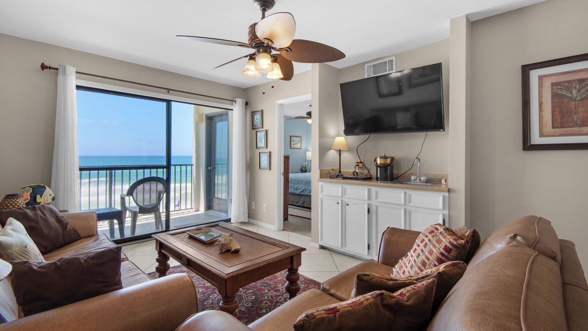 Een ruime woonkamer met balkon en uitzicht op zee, een flatscreen-tv boven de keukenbar, en een plafondventilator.