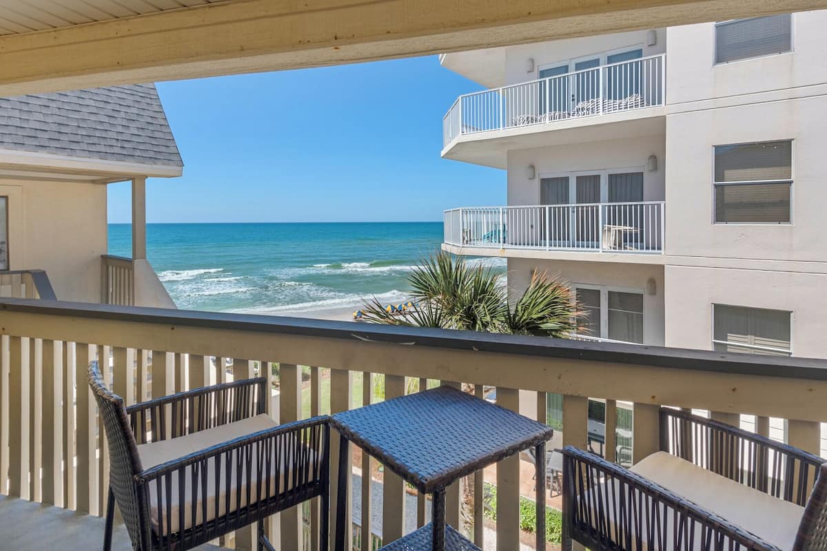 Een balkon met rieten meubilair met uitzicht op een strand en aangrenzende gebouwen.