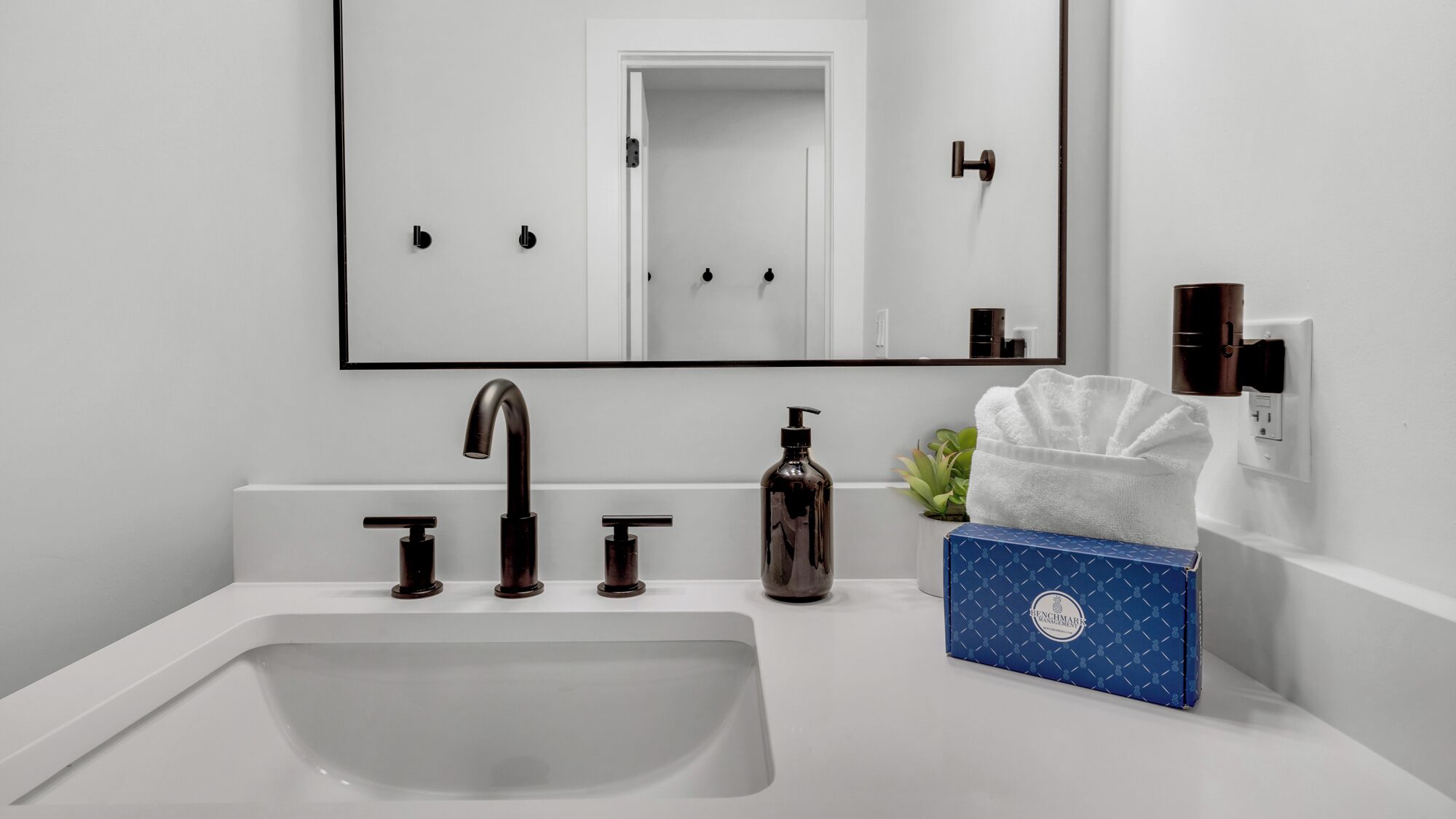 A modern bathroom with a white sink, black faucets, a mirror, and a blue box with a logo.