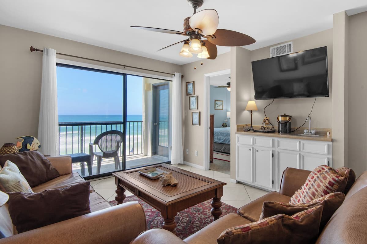 A spacious living room with a balcony overlooking the ocean, a flat-screen TV mounted above the kitchen counter, and a ceiling fan.