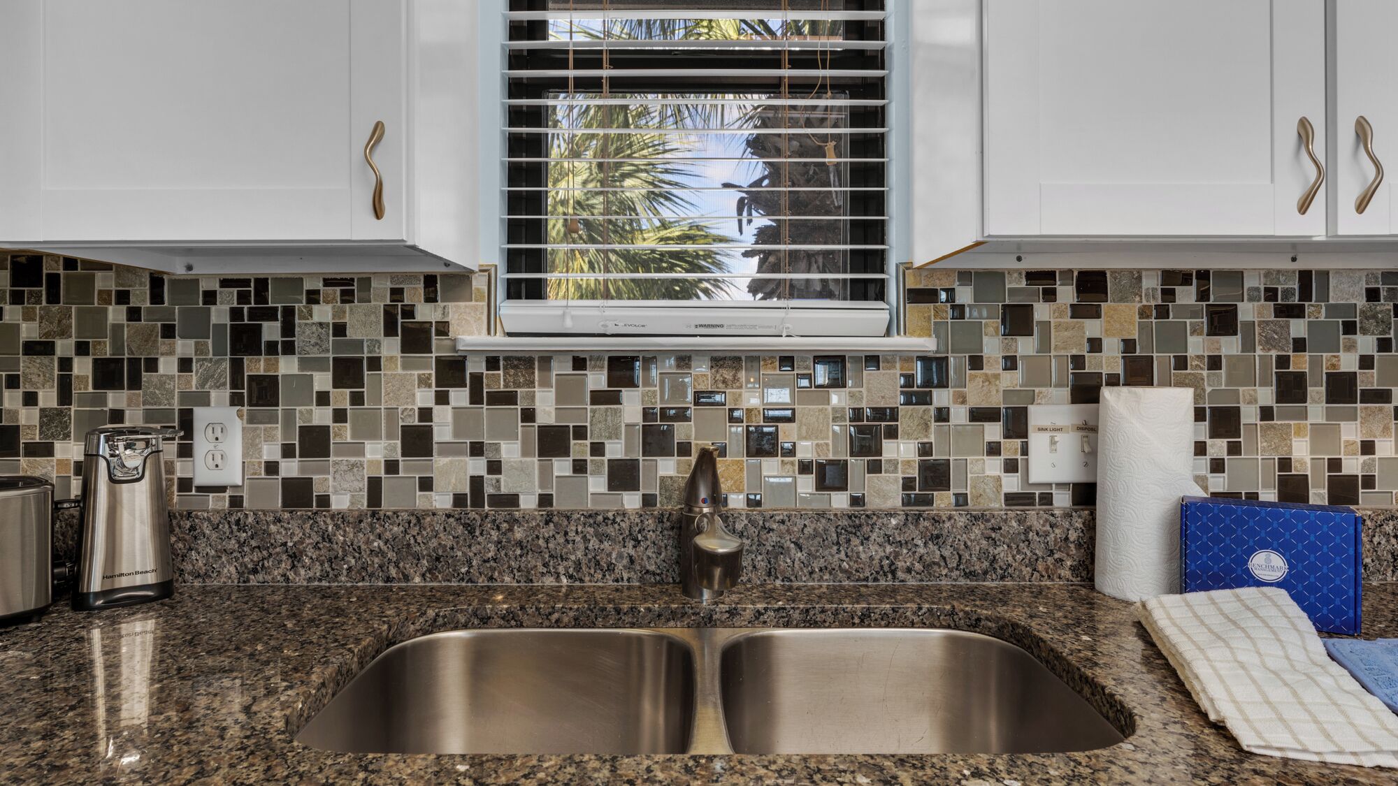 A kitchen with a double sink, granite countertop, and a window with blinds.