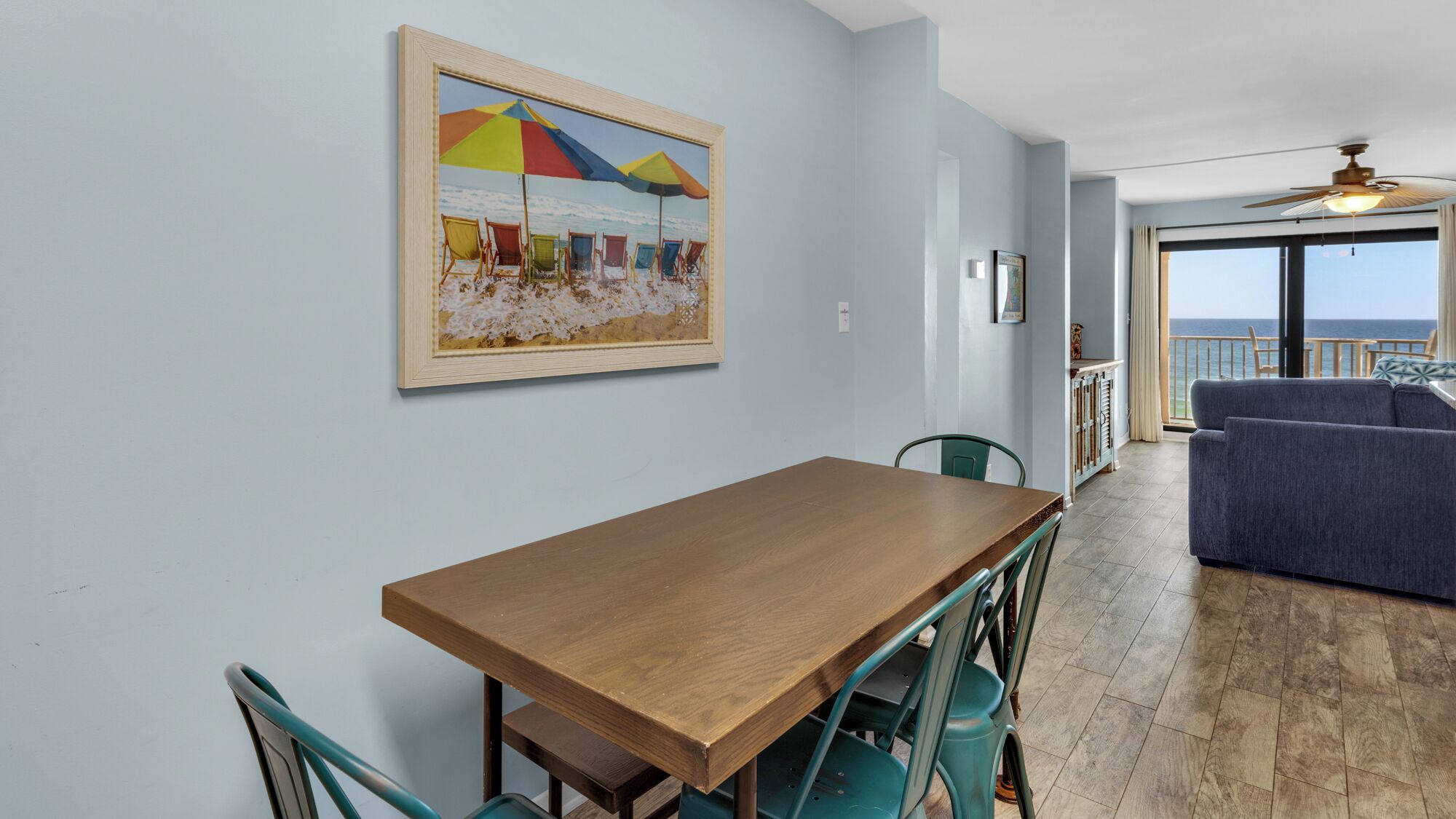 A dining area with a wooden table and teal chairs, a colorful beach-themed painting on the wall, and a view of the ocean through large windows.