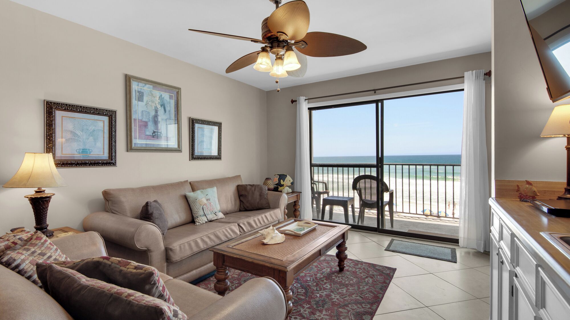 A living room with a sofa, armchairs, a coffee table, and a ceiling fan. There are framed pictures on the wall and a view of the beach through a large sliding glass door.