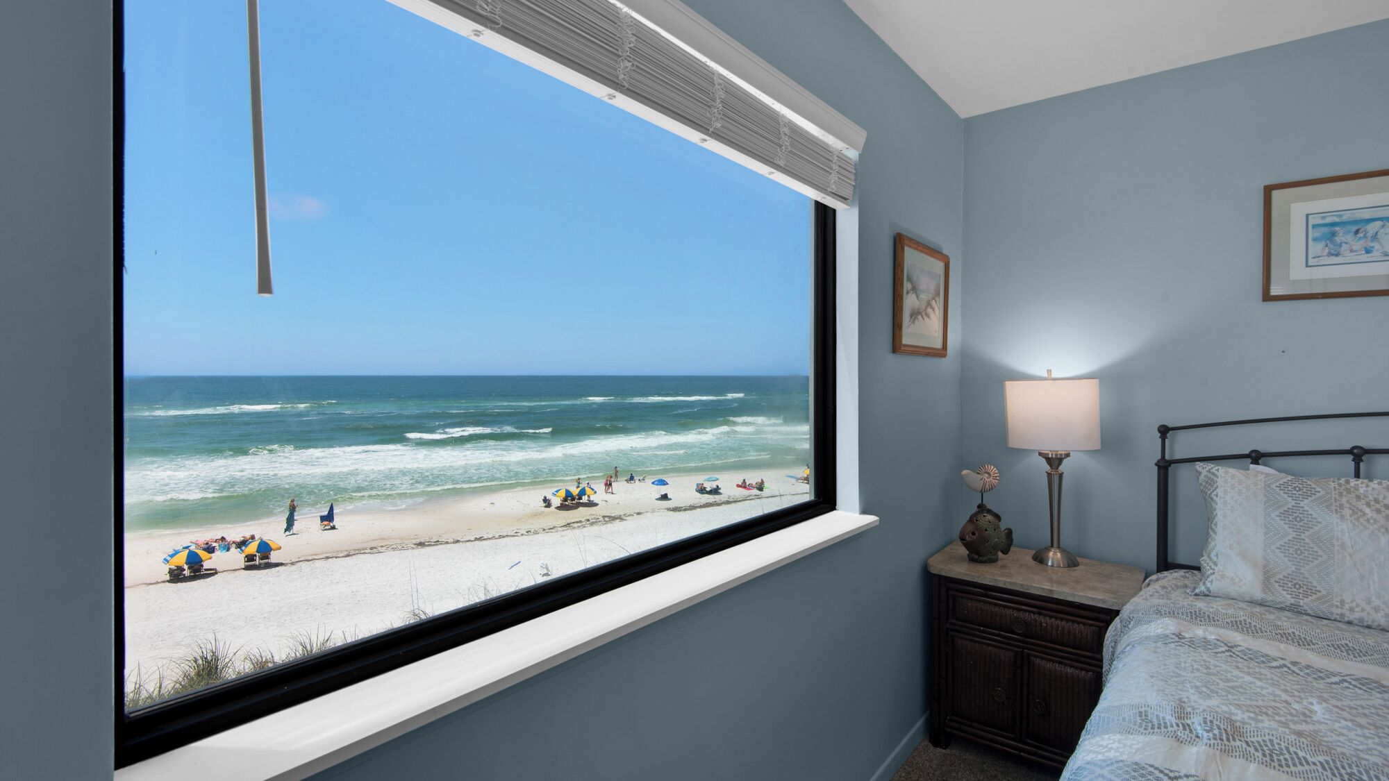 A bedroom with a large window overlooking a beach, a bedside table with a lamp, and a bed with patterned bedding.