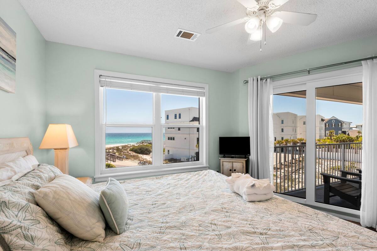 A bedroom with a bed, a TV, a window with a view of the beach, and a balcony with a railing.