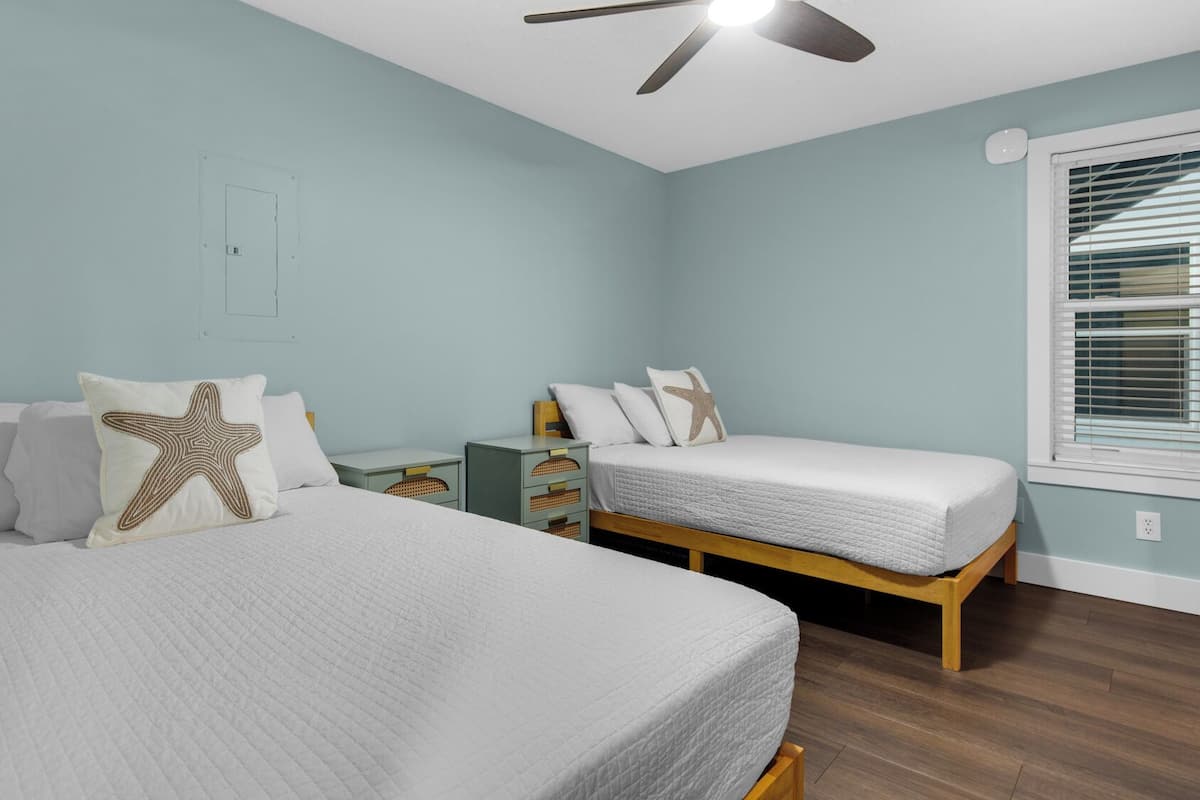 A bedroom with two beds, a ceiling fan, a window with blinds, and a wall socket.