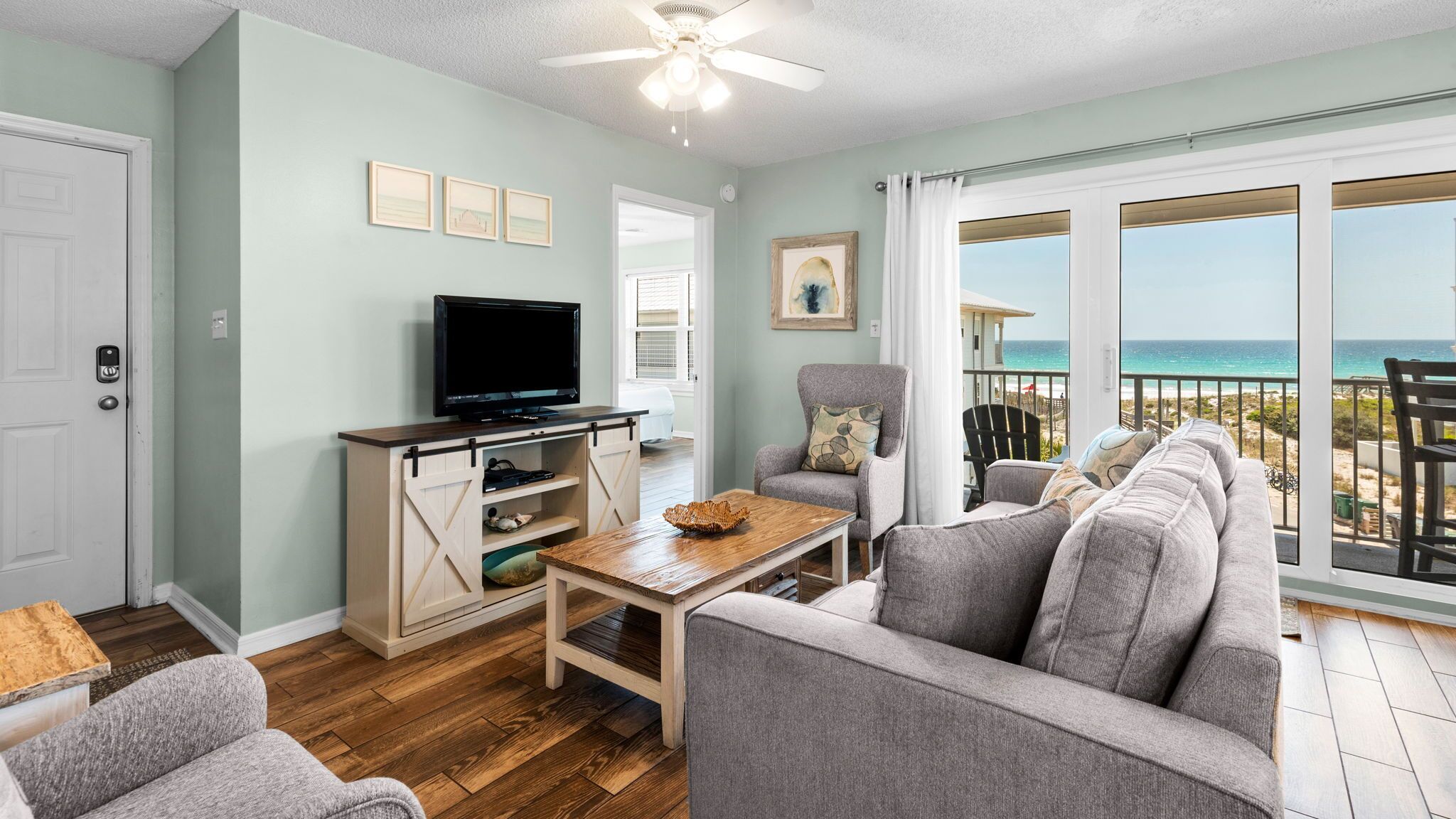 A living room with a TV, a sofa, a chair, and a table. There is a balcony with a view of the ocean.