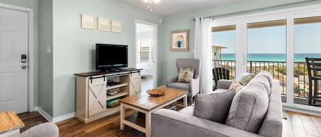 A living room with a TV, a sofa, a chair, and a table. There is a balcony with a view of the ocean.