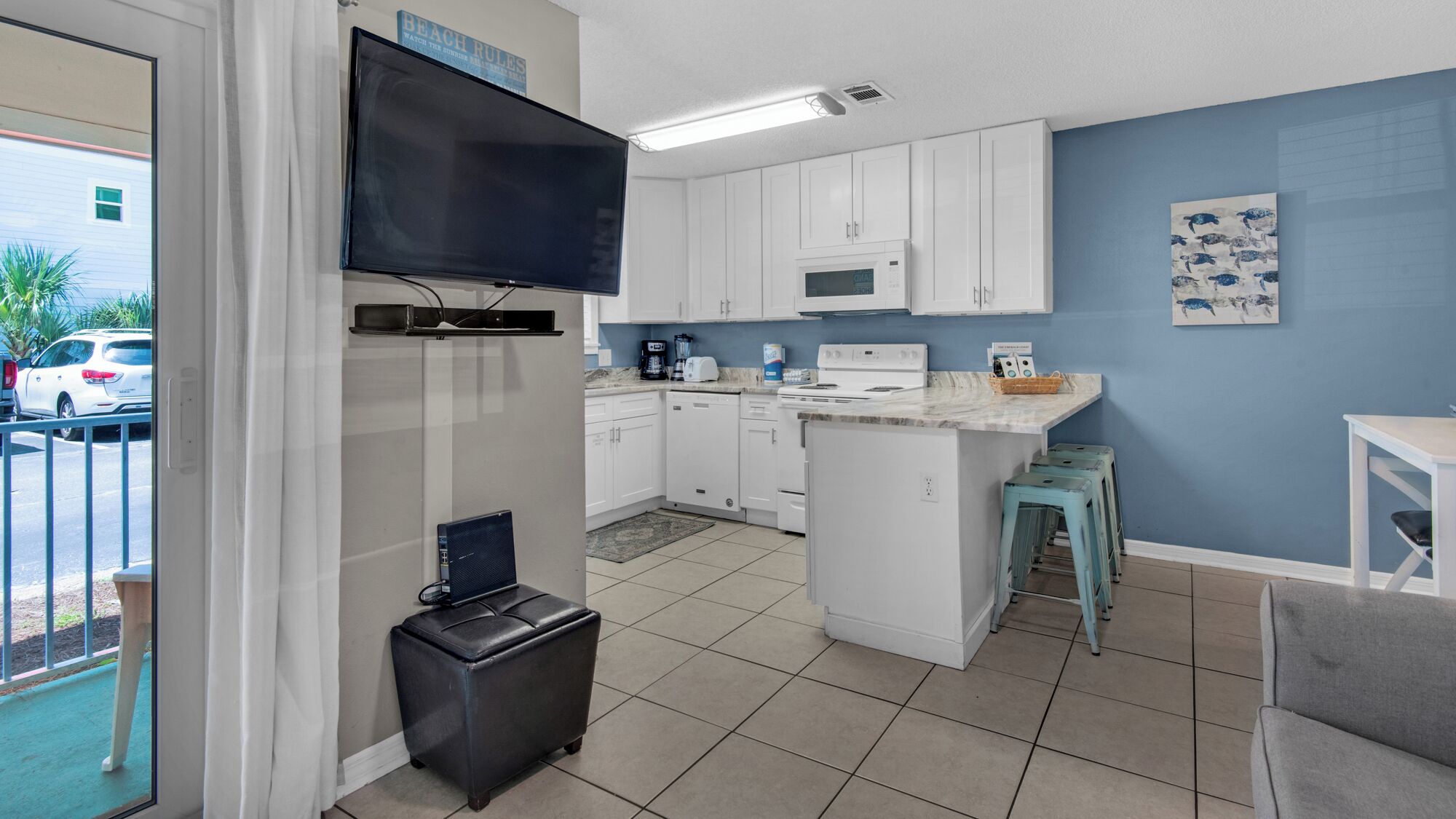A compact kitchen with white cabinets, a microwave, and a small island. There is a television mounted above the counter and a painting on the wall.
