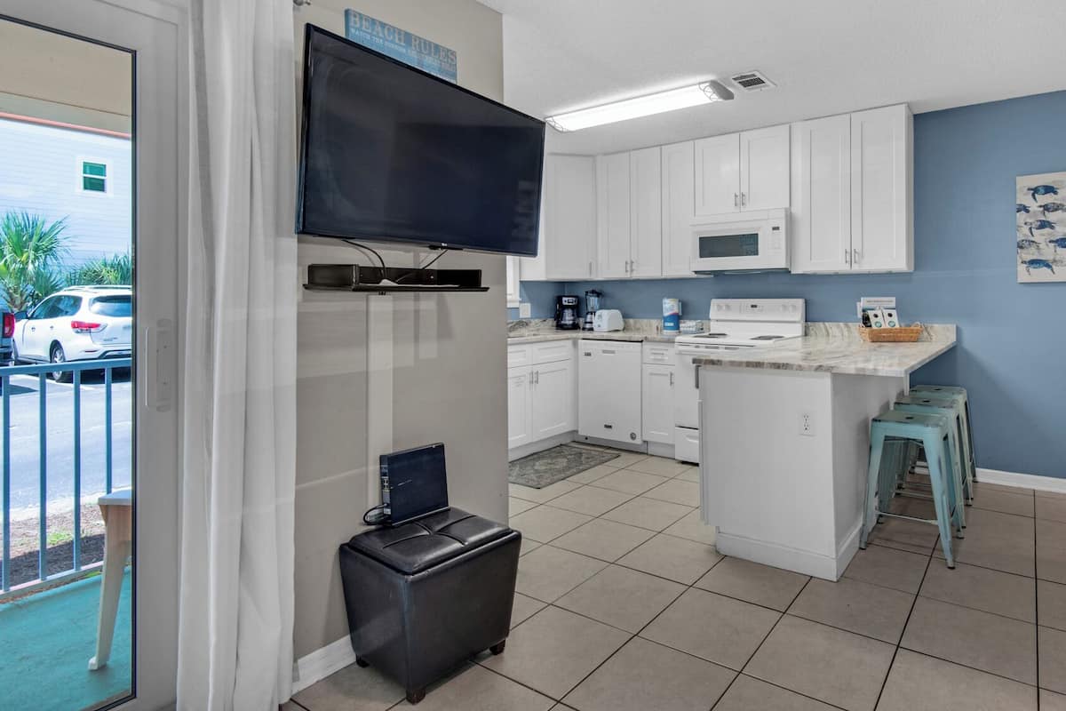 A compact kitchen with white cabinets, a microwave, and a small island. There is a television mounted above the counter and a painting on the wall.