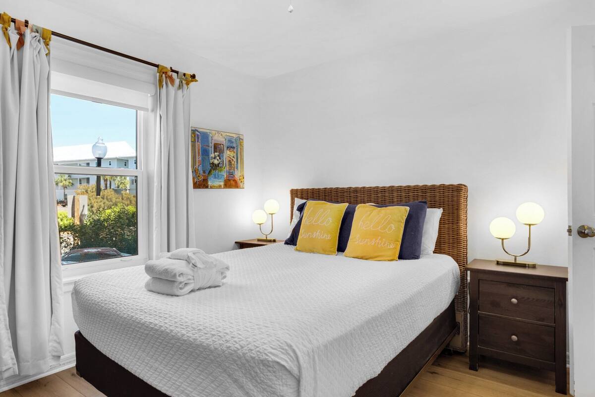 A neatly made bed with a white bedspread and two pillows, a window with white curtains, a wall-mounted painting, and a bedside table with a lamp.