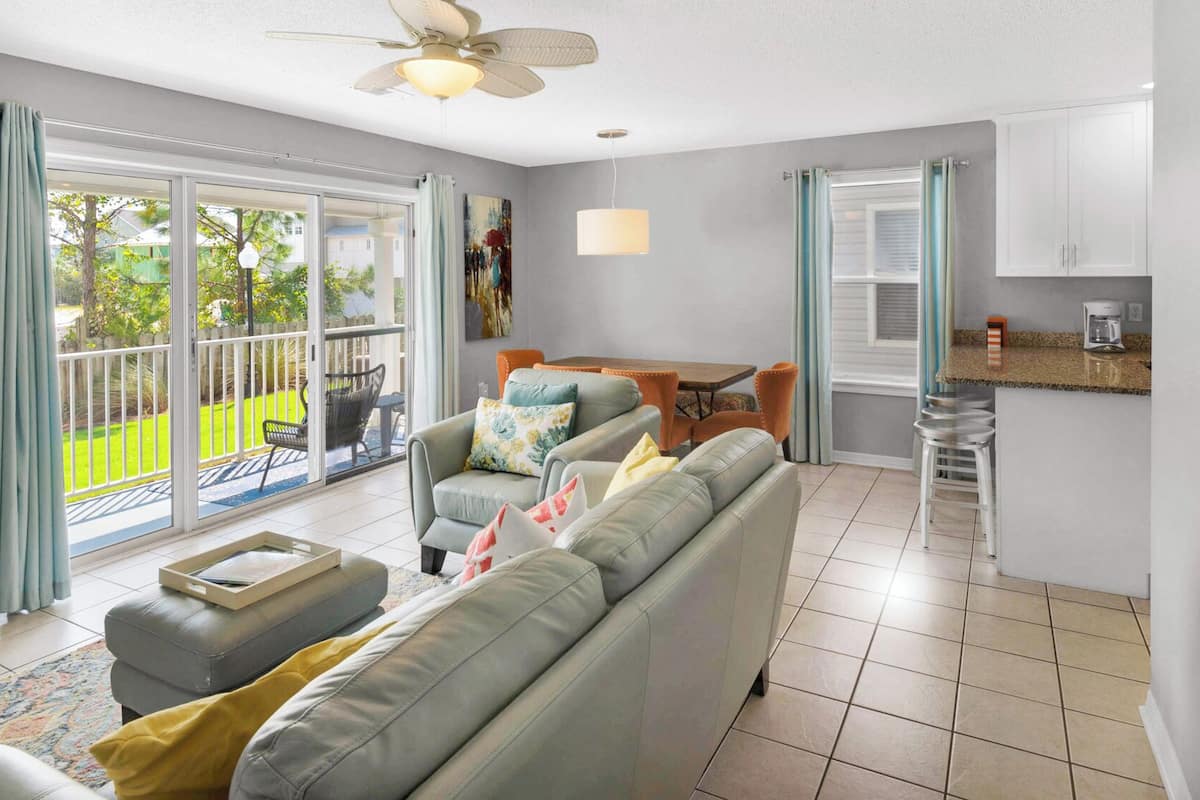 A modern living room with a grey sofa, a coffee table, and a dining area with a table and chairs. There is a kitchen area with a countertop and a coffee machine.