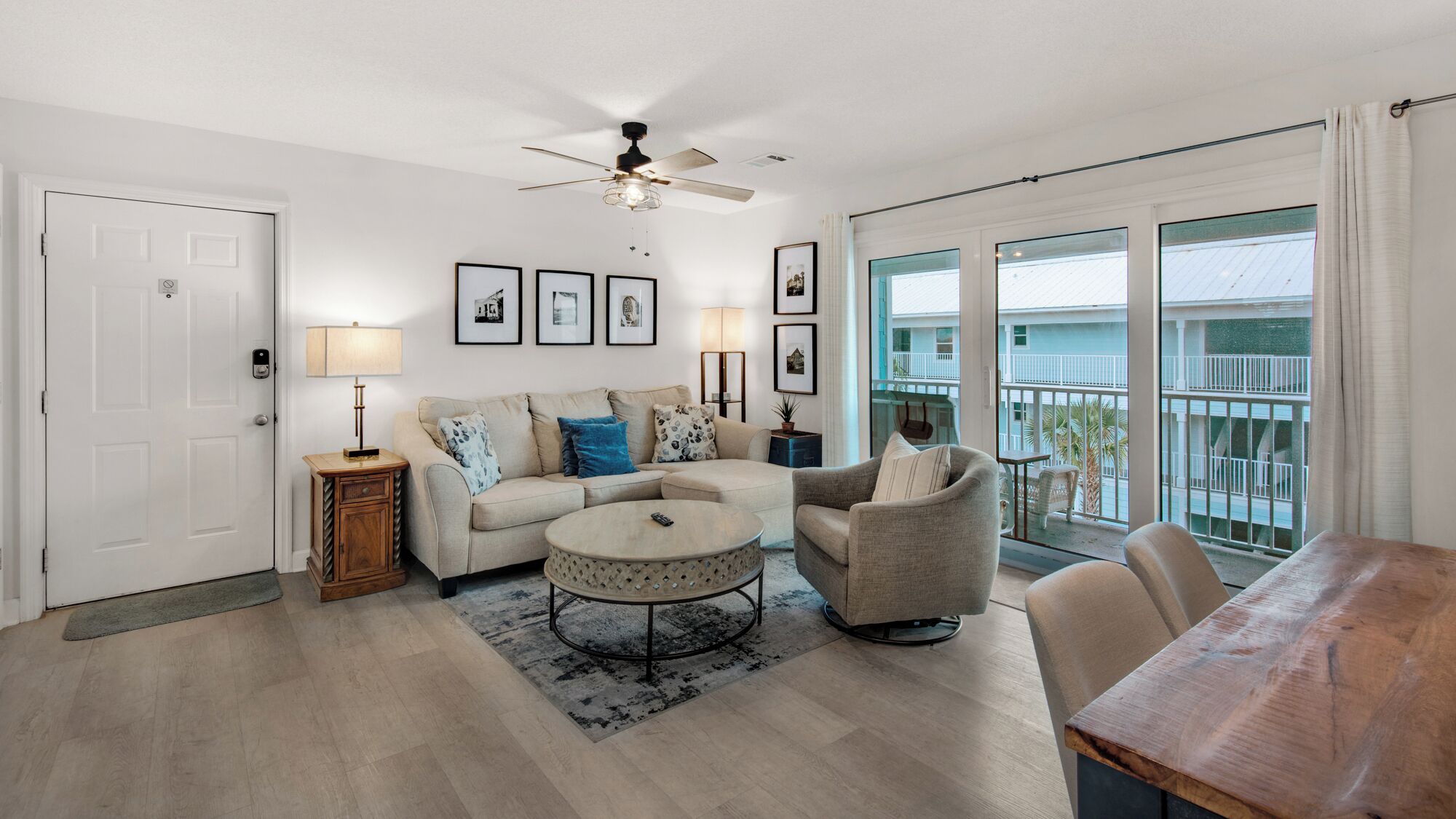 A spacious living room with a beige sofa, a round coffee table, and a gray armchair. There is a ceiling fan, a door with a doorknob, and a window with a view of a balcony.
