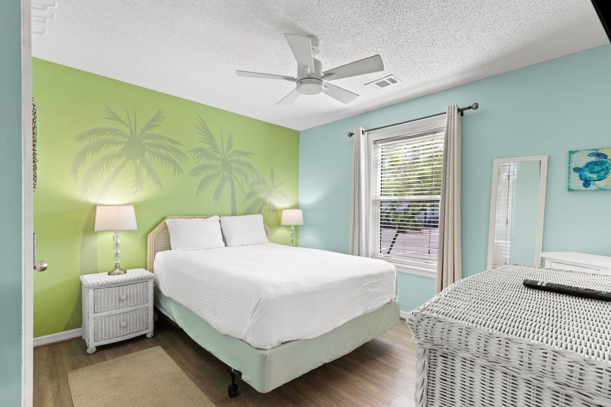 A bedroom with a bed, nightstands, a ceiling fan, a window with blinds, and a painting on the wall.
