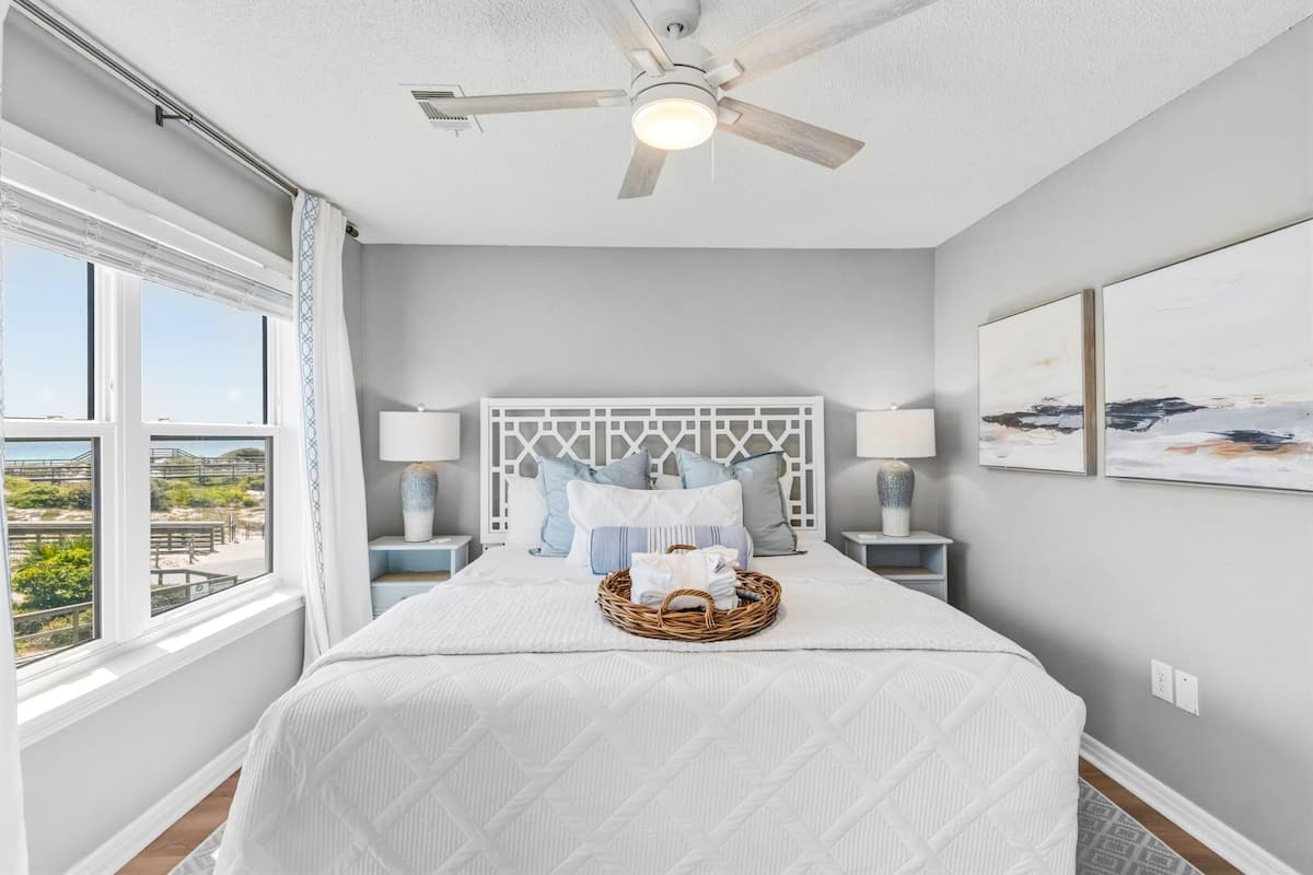 A bedroom with a bed, a ceiling fan, bedside lamps, a window with a view, and artwork on the wall.